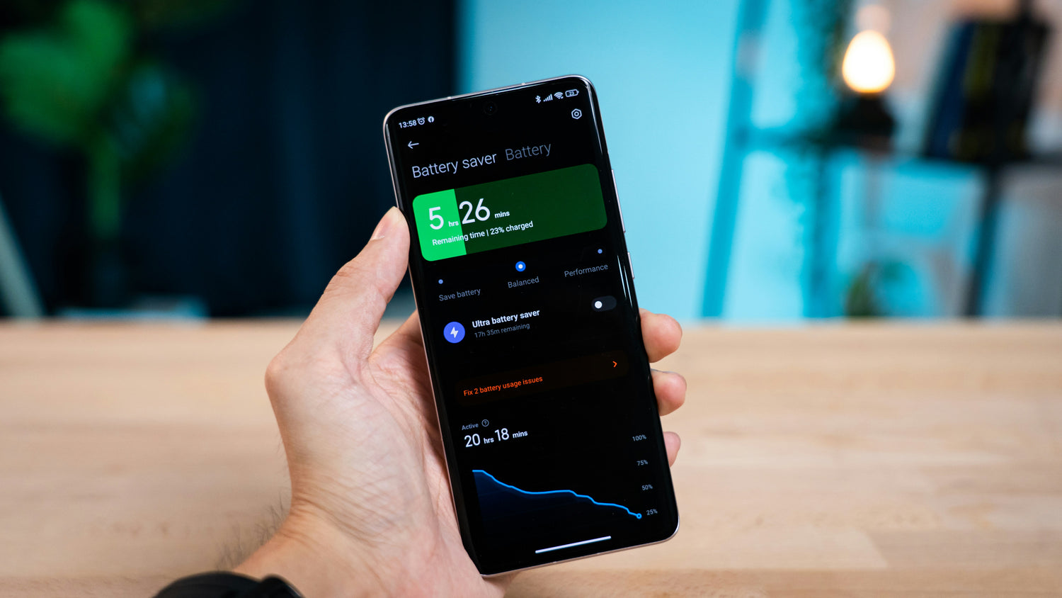 A hand holding a smartphone displaying a electric vehicle battery optimization app interface, with a softly blurred indoor background showing a wooden table and blue toned decor.