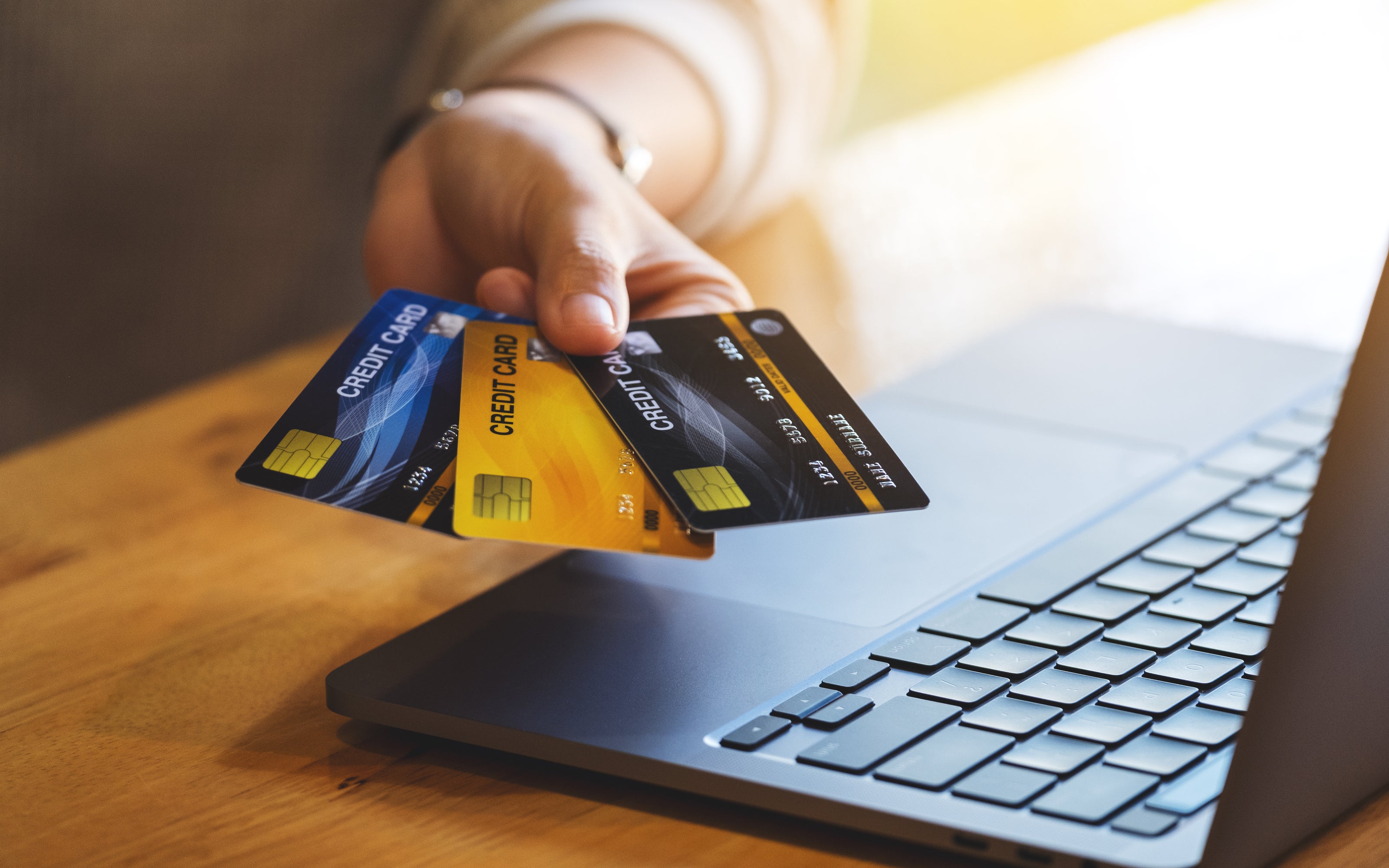 Hand holding multiple credit cards above a laptop on a wooden surface