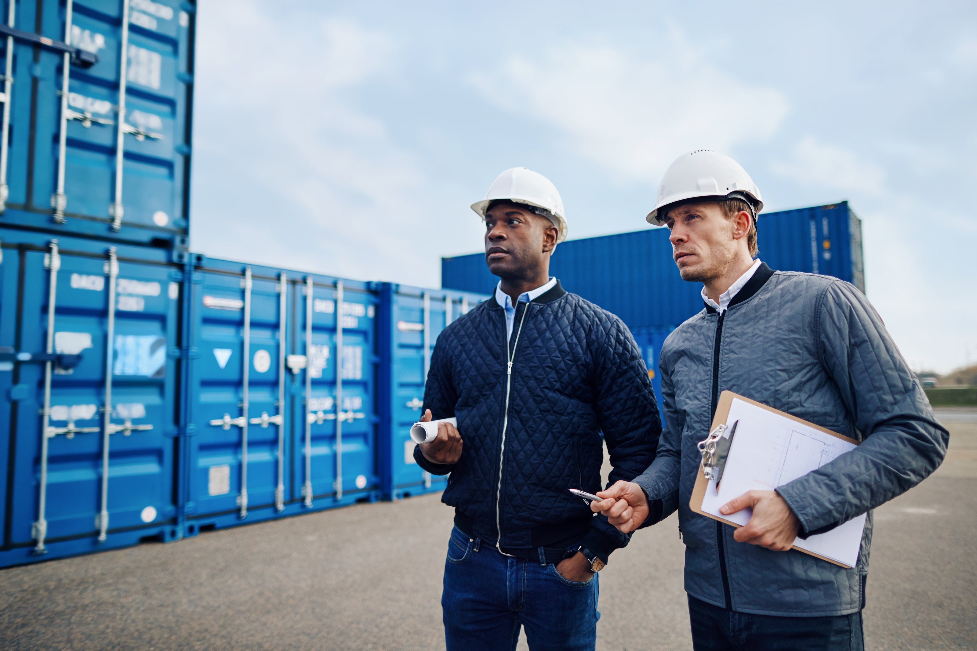 Two men in hard hats standing in front of blue shipping containers.