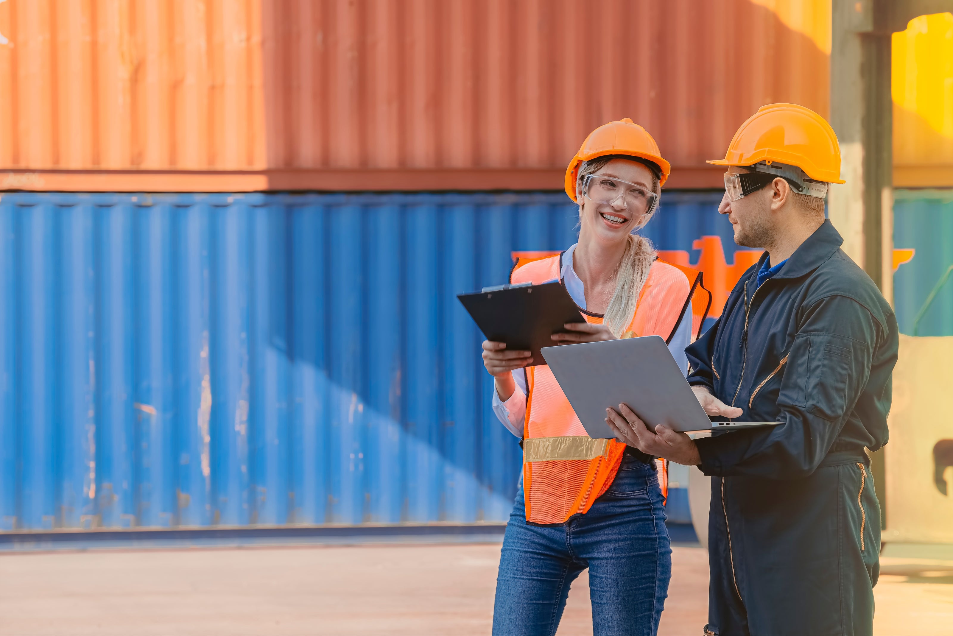 Two workers in hard hats and safety vests using laptops in front of shipping containers.