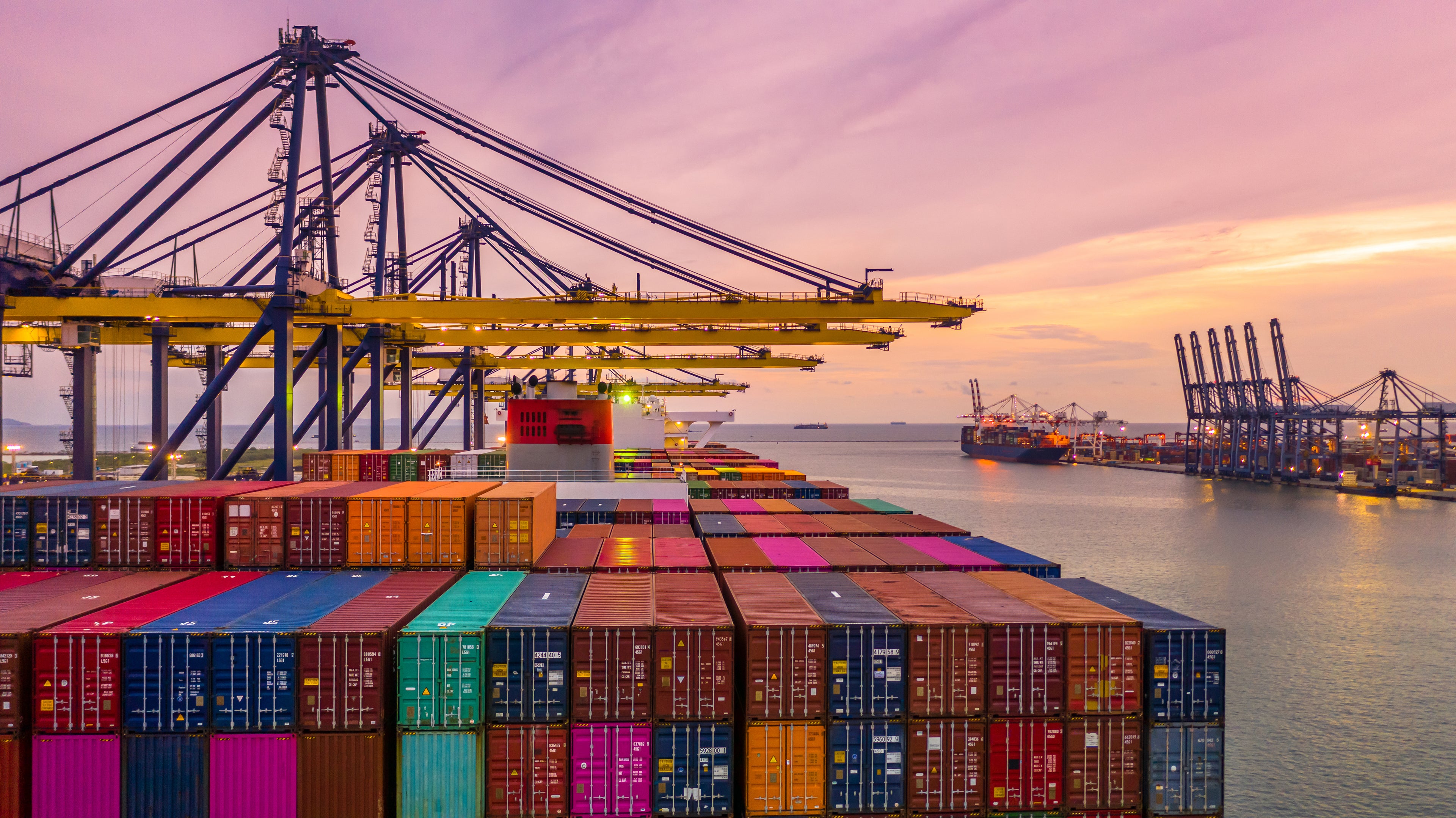Container ship at a port with colorful containers and cranes against a sunset sky.