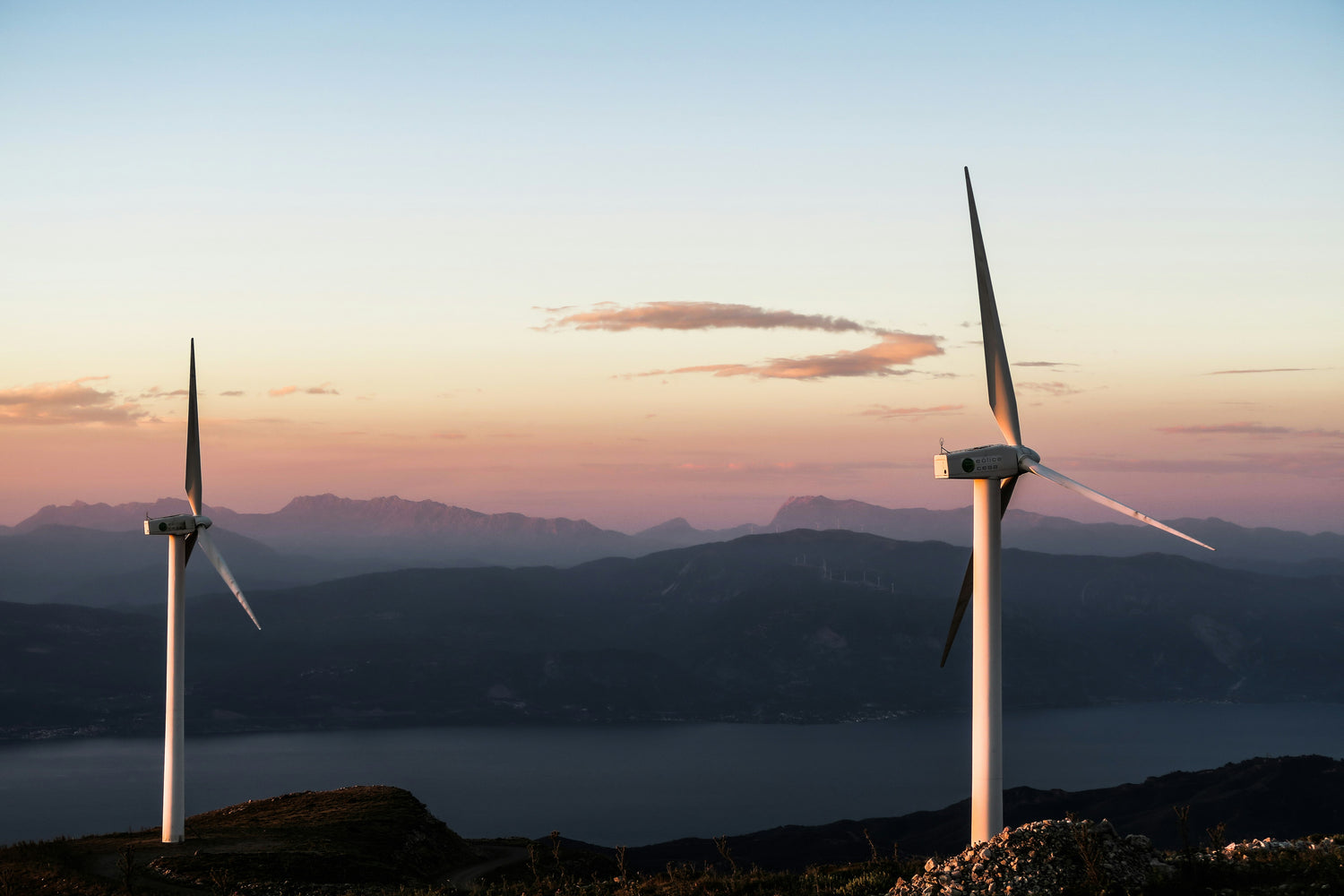 Two large wind turbines stand on a mountainside overlooking a lake and distant mountain ranges at sunset, with a pale blue sky and soft orange clouds. EV energy management displaying power consumption, charging loads, and energy optimization metrics.