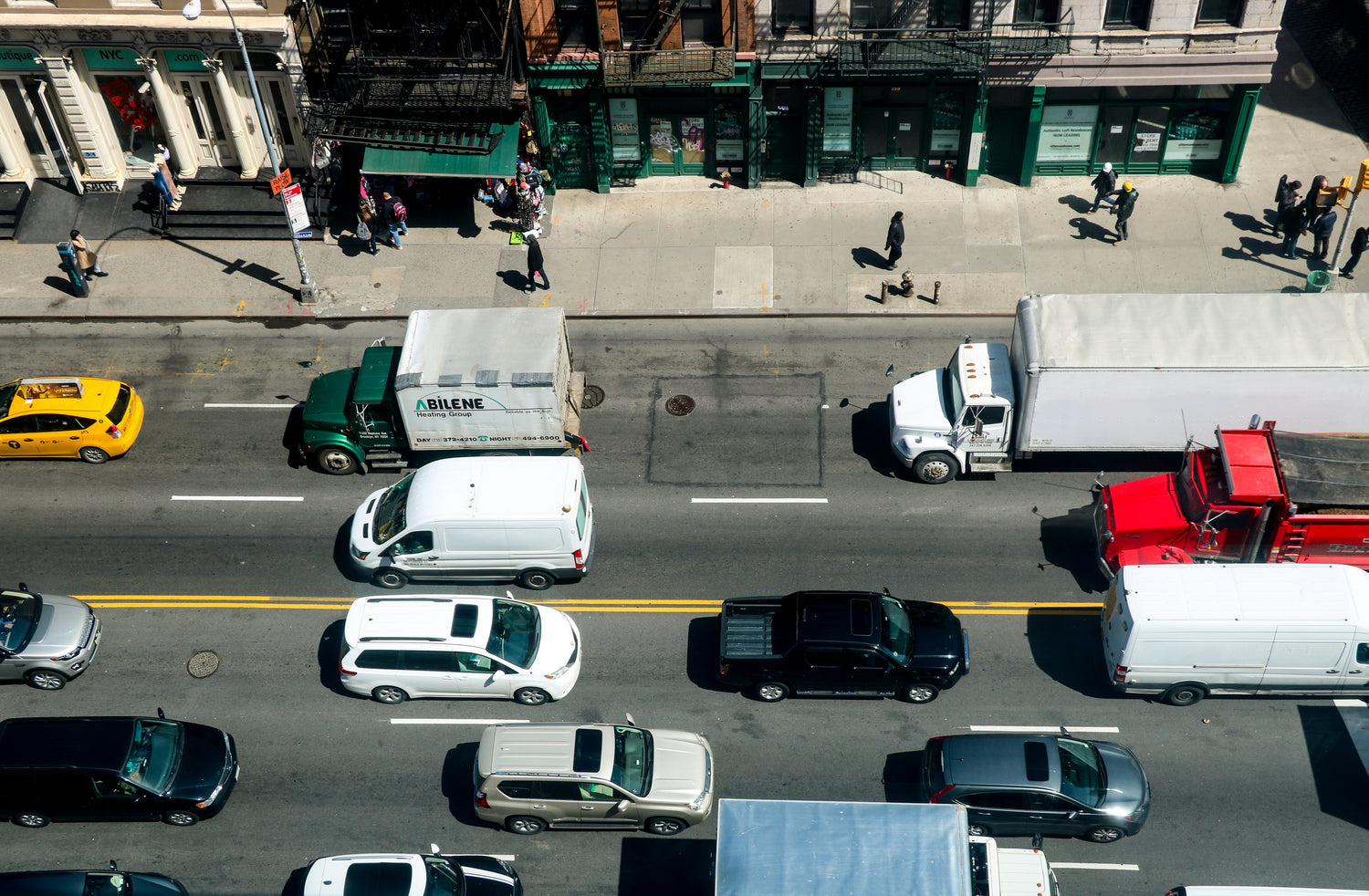Aerial view of a busy city street with multiple cars, trucks, and delivery vehicles moving along the road, with pedestrians and storefronts visible along the sidewalk.