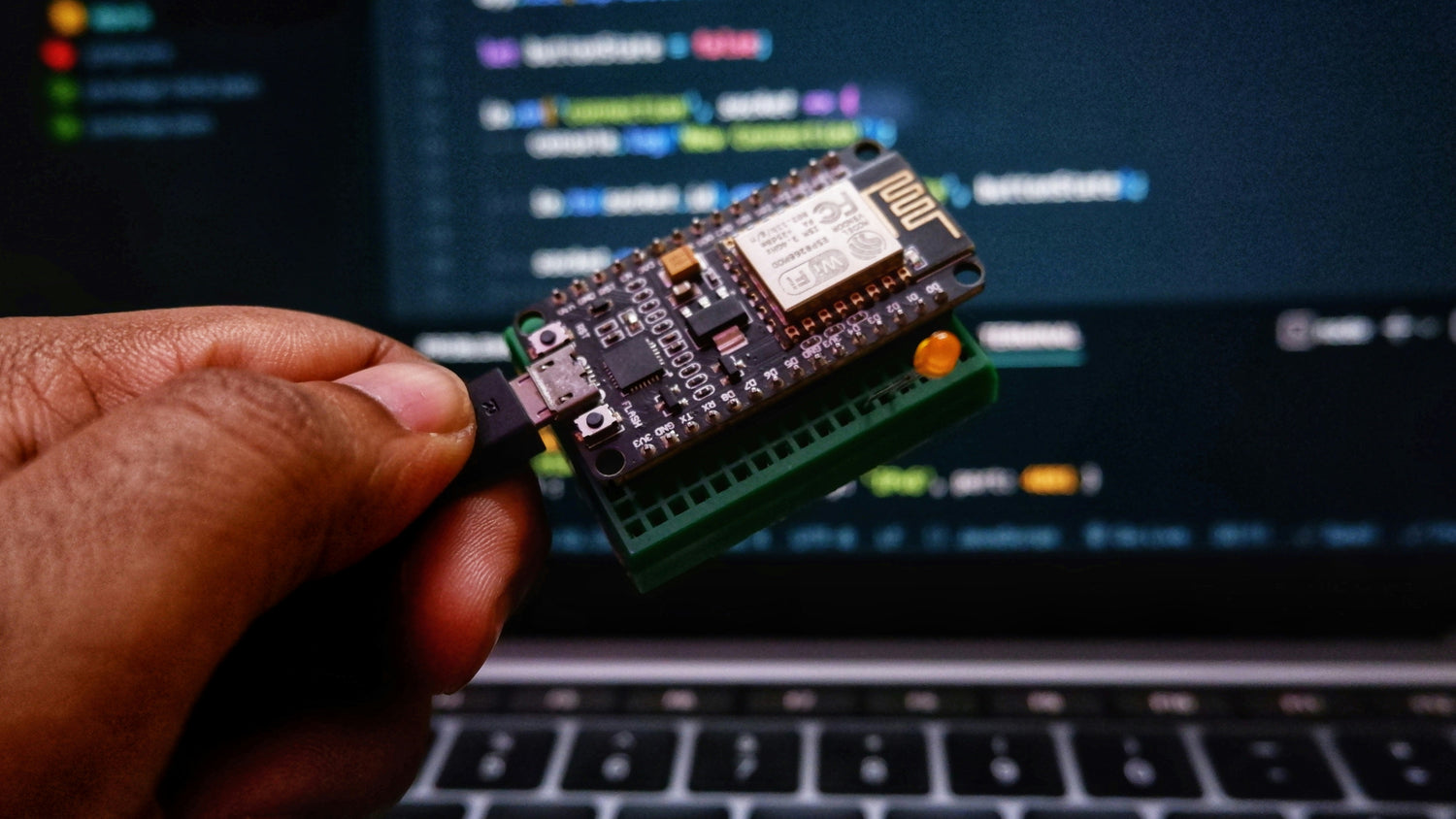 A hand holding a small electronic circuit board with a green breadboard connector and a wireless module, positioned in front of a blurred laptop screen. Connected electric vehicles, charging stations, and IoT devices exchanging real-time operational data.